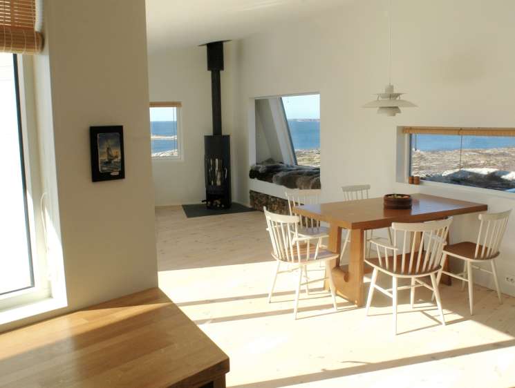 Modern cabin interior with wooden dining table, white chairs, black wood stove, and ocean views through large windows.
