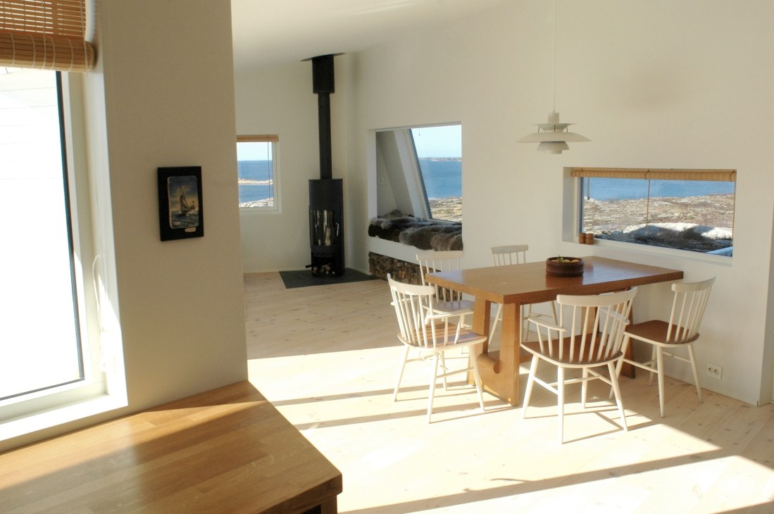 Modern cabin interior with wooden dining table, white chairs, black wood stove, and ocean views through large windows.