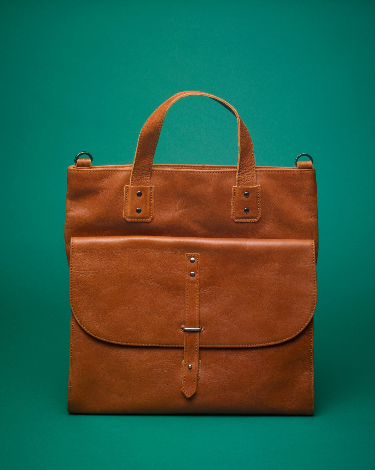 Tan full-grain leather Courier bag with double handles and front pouch against teal background.