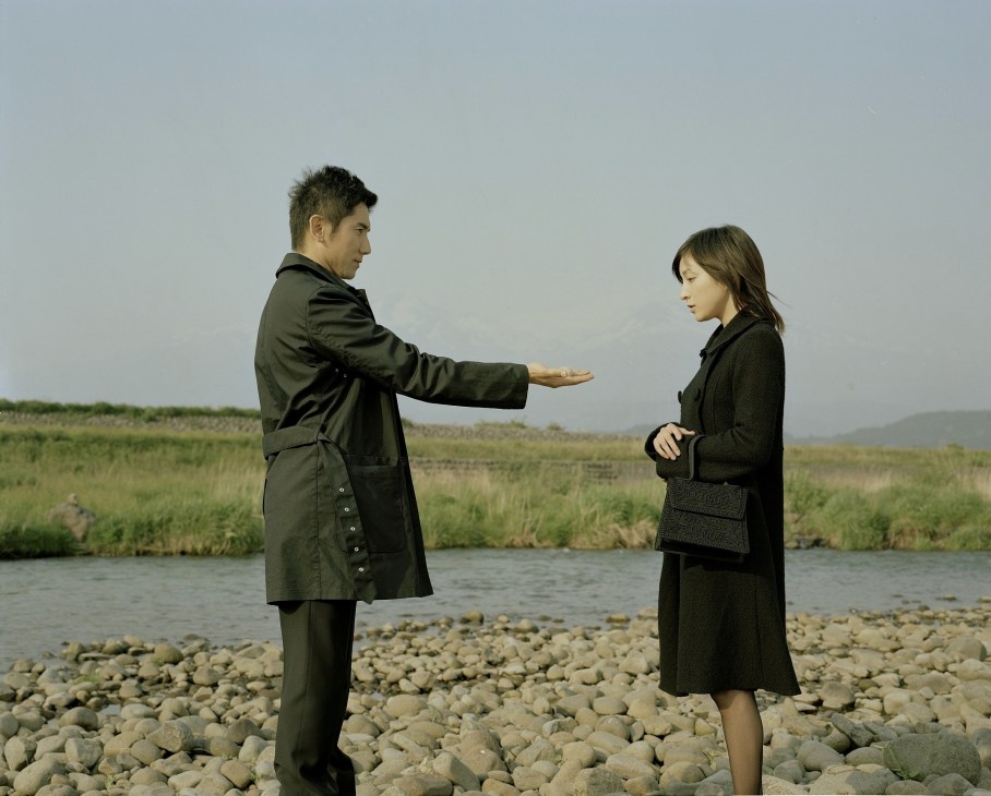 Two figures in dark coats stand on a rocky riverbank in a misty landscape.