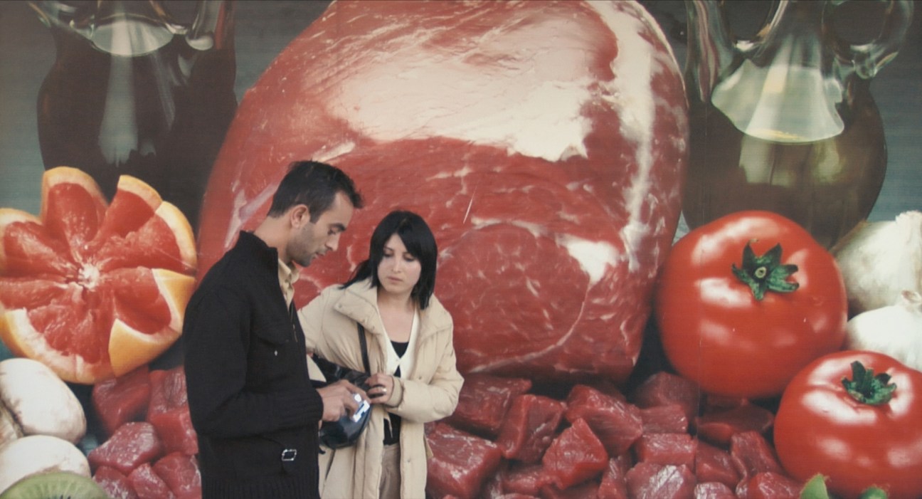Two figures standing in front of a large-scale mural depicting raw meat, tomatoes, and grapefruit.
