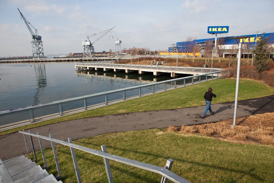 Person jogging along waterfront path at Erie Basin with IKEA store and industrial cranes visible across the water.