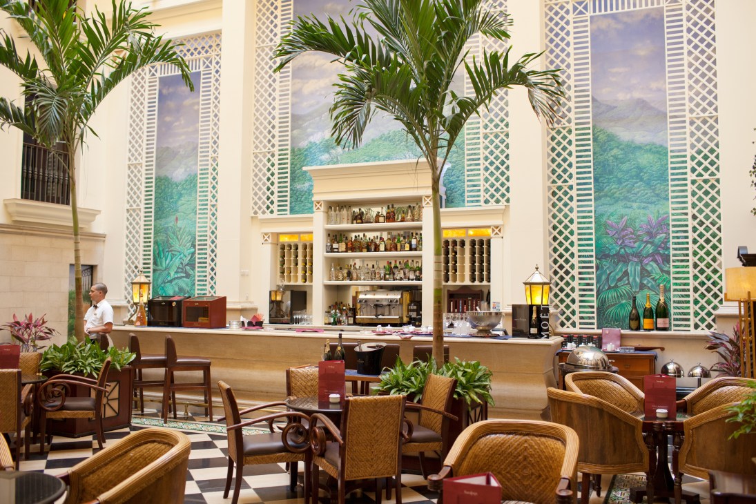 Interior of Hotel Saratoga bar with tropical plants, wicker furniture, and art deco-style wall panels.