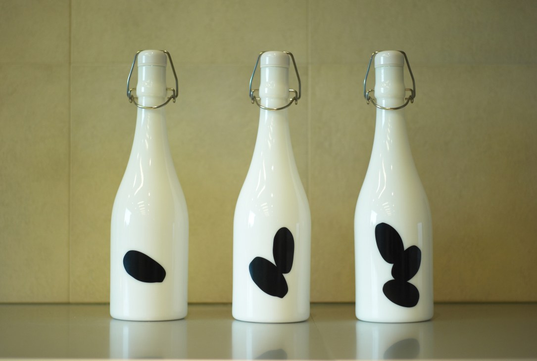 Three white sake bottles with wire flip-top closures displaying black rice-kernel silhouettes.