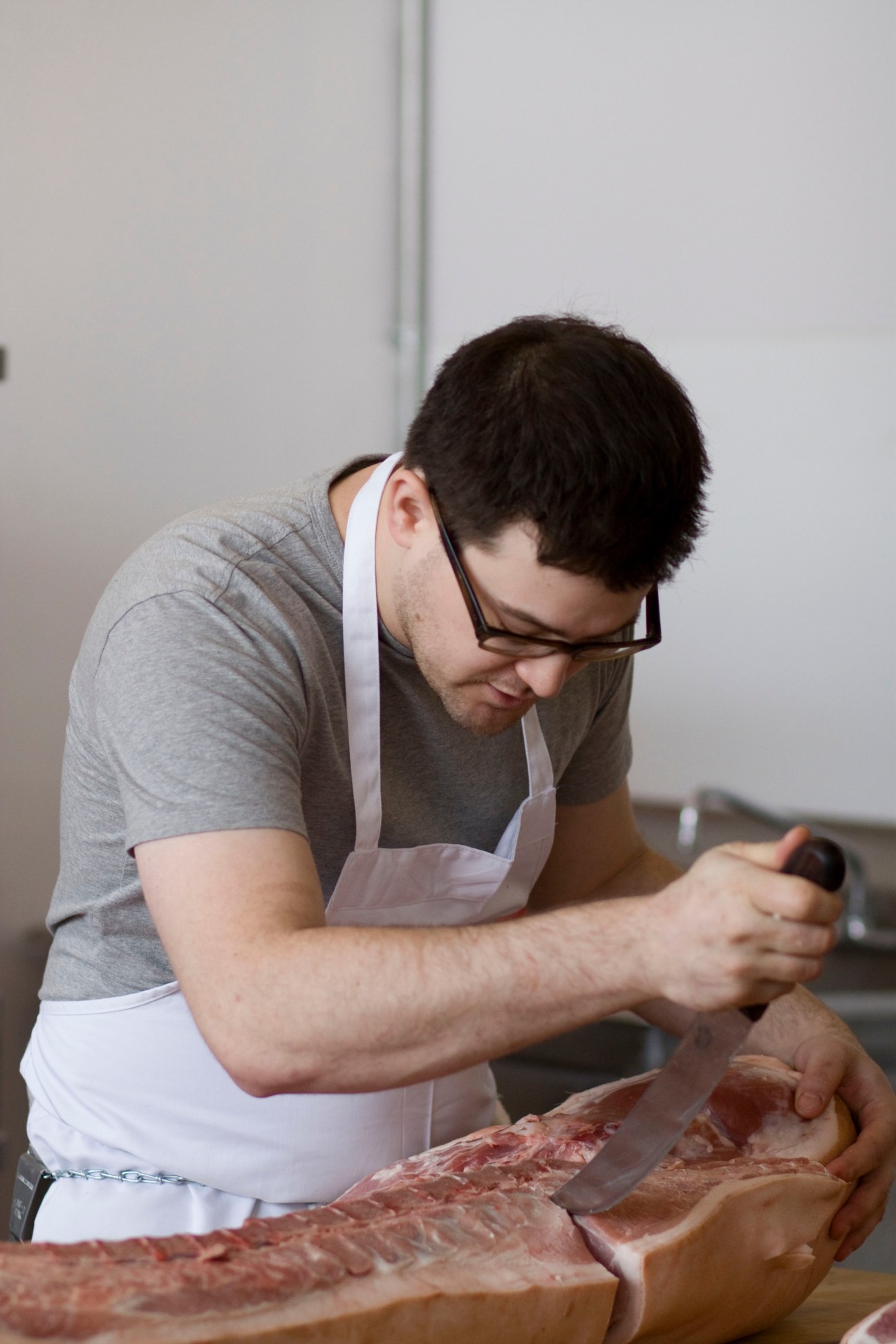 Butcher in white apron and glasses carefully slicing raw meat on a cutting board.