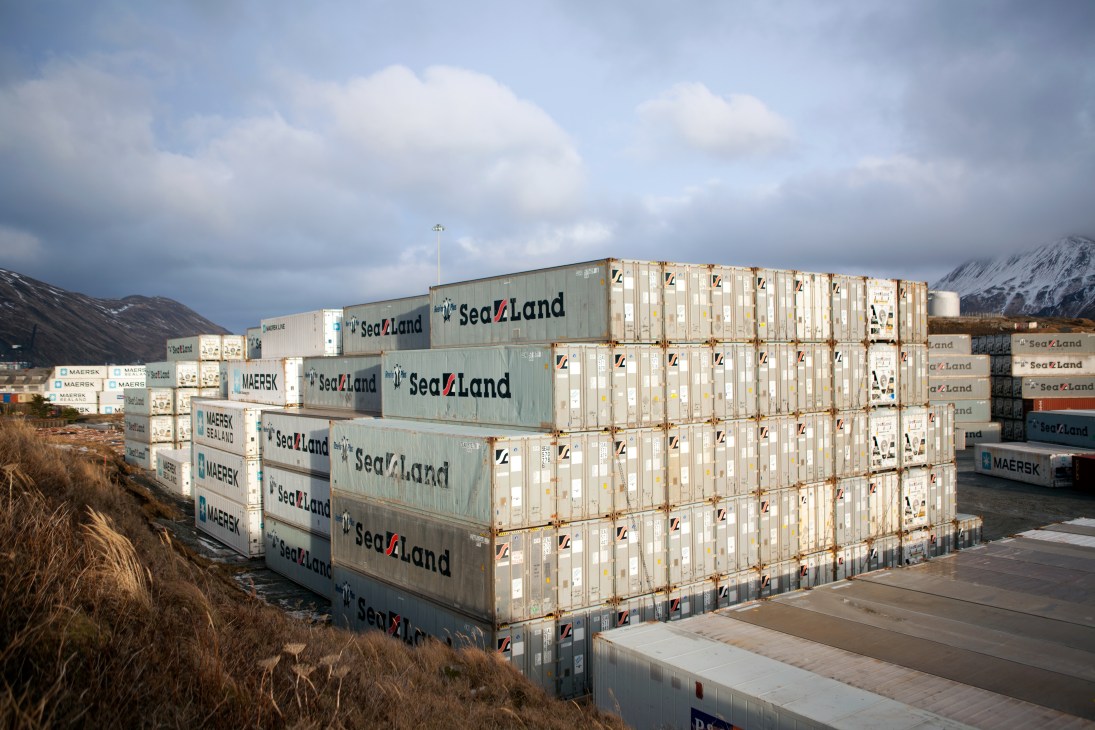 Stacked shipping containers labeled SeaLand and Maersk at Dutch Harbor port with snow-capped mountains in background.