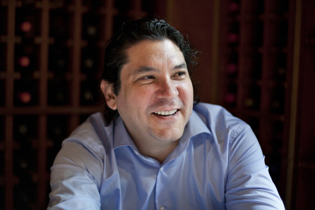 Gastón Acurio, a Peruvian chef in a blue shirt, smiles at the camera in front of a wine rack.
