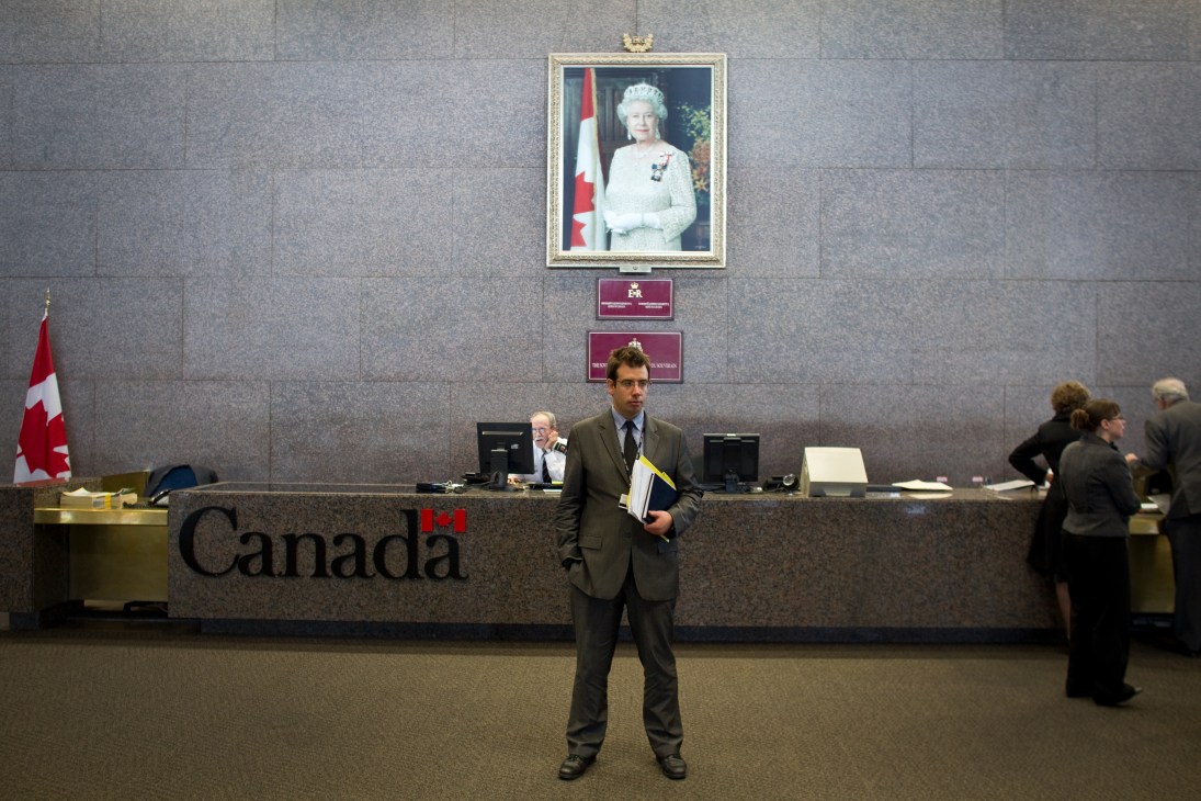 Interior of Canadian government office with man in suit holding documents standing below Queen Elizabeth II portrait.