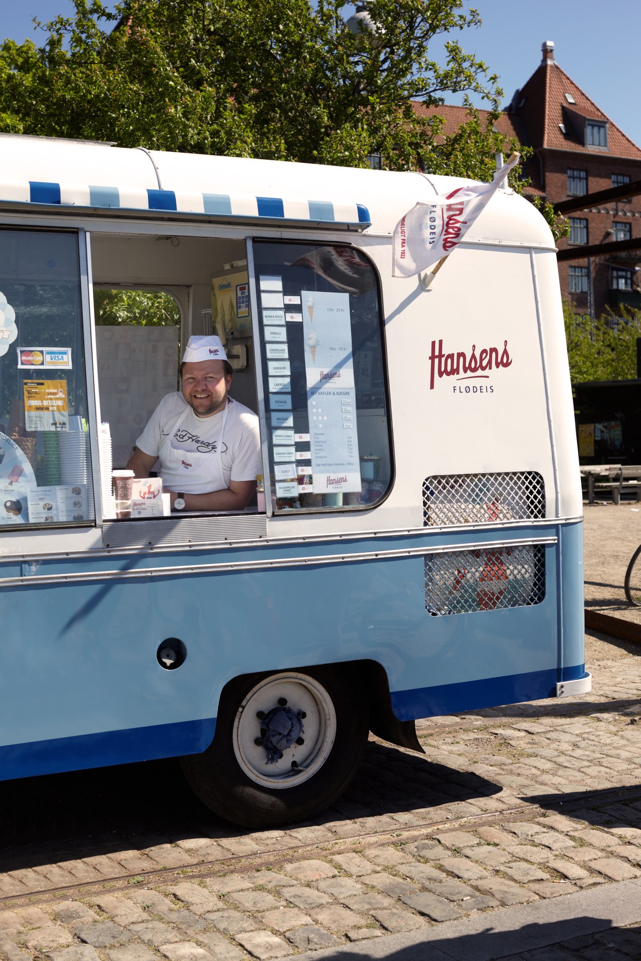 Man serving ice cream from a blue and white vintage Hansens Flodeis food cart on a cobblestone street.