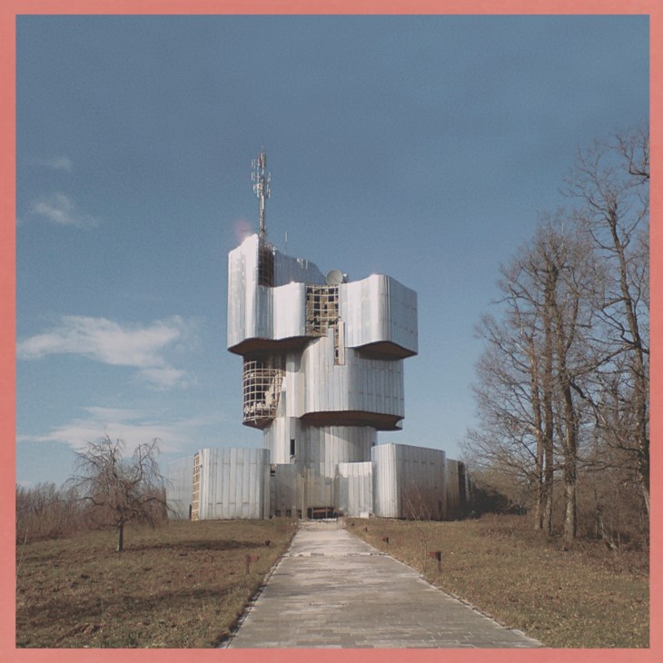 Modernist white tower building with protruding balconies and communication antenna in sparse landscape.