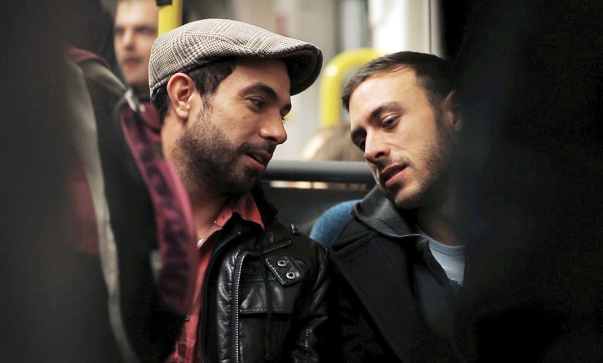 Two men conversing in a subway car; one wears a flat cap and plaid shirt, the other a blue shirt.