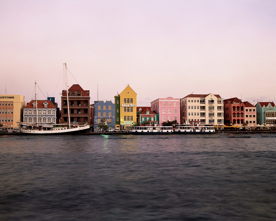 Colorful colonial buildings line the waterfront of Willemstad's St Annabaai channel with boats moored below.