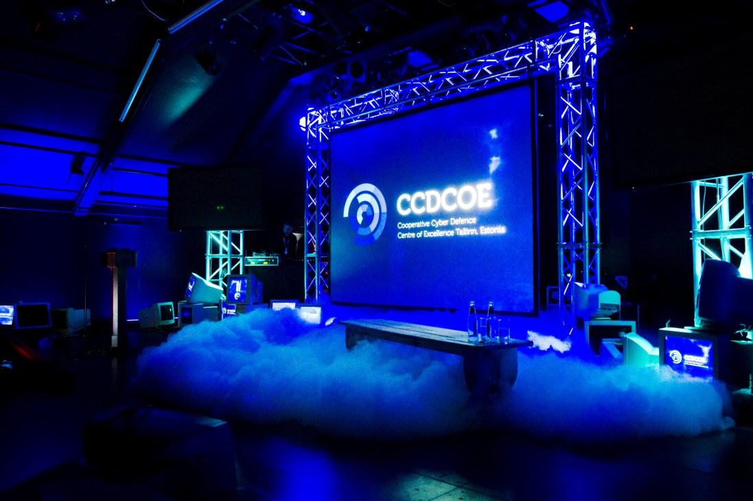 A conference stage with blue lighting displaying the CCDCOE logo and text, surrounded by metal truss framework.