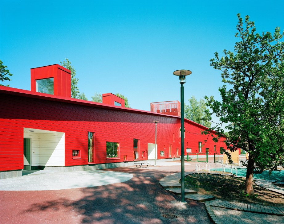 Red timber-slatted daycare centre in Espoo, Finland with glass panel towers on roof and playground area.