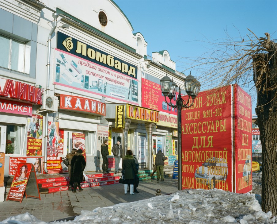 Street market in Blagoveshchensk with colorful red storefronts and signage in Russian and Cyrillic text.