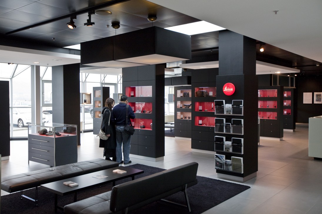Interior of a modern Leica camera showroom with black display cases, red shelving, and customers browsing.