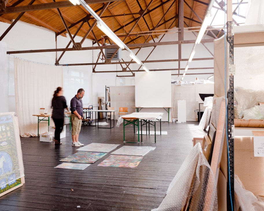 Two people working in a large industrial space with wooden beams, organizing artworks and furniture for an exhibition.