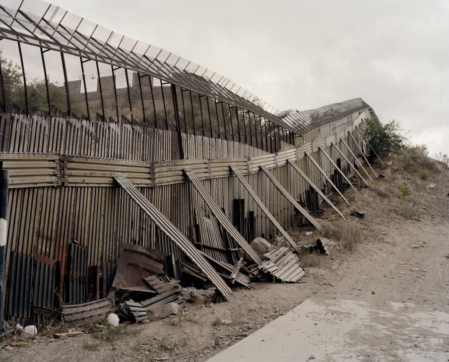 Metal border fence on US side with damaged and deteriorating sections along dirt terrain.