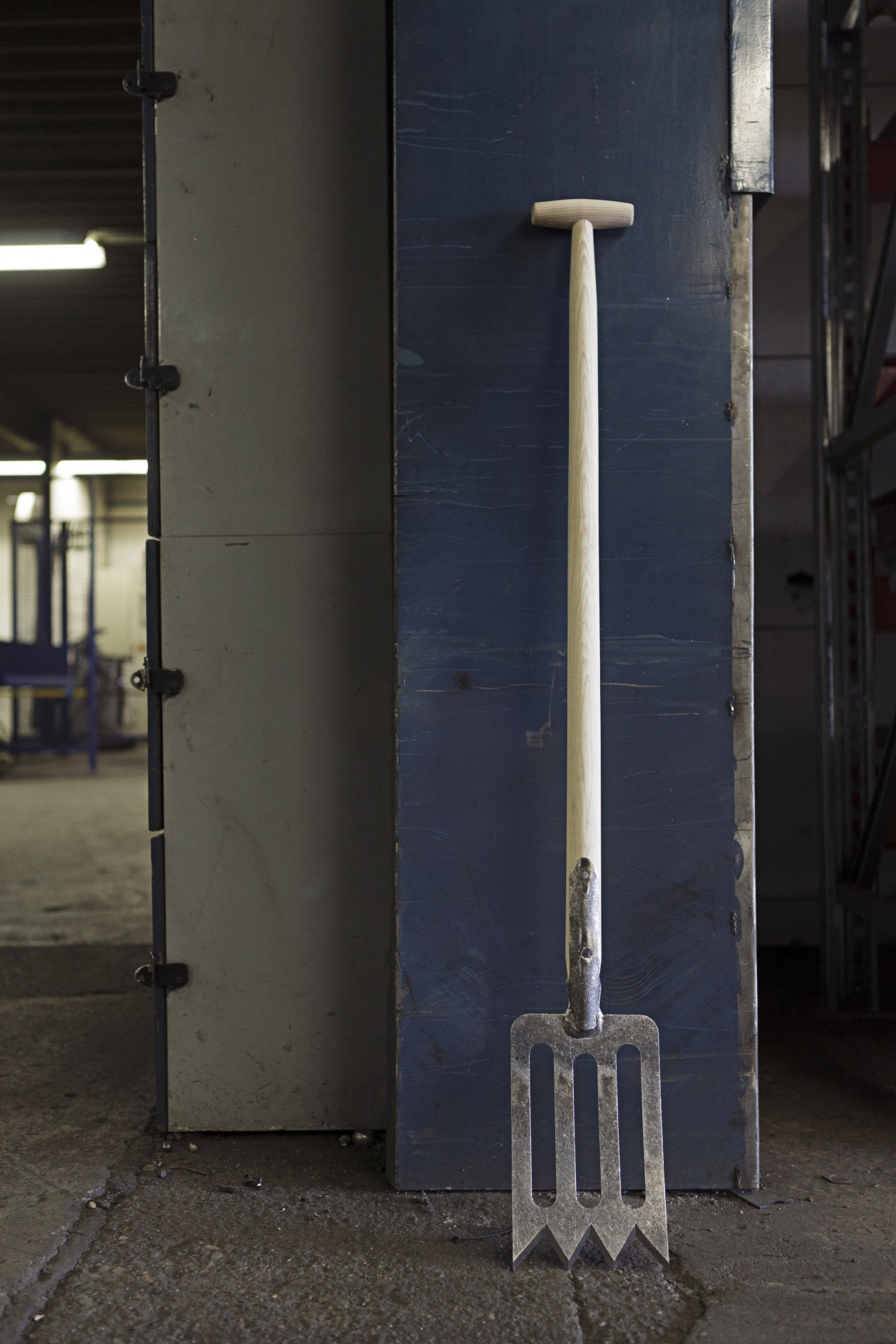 DeWit spork garden tool with metal fork-like head and wooden handle leaning against dark blue panel in factory.