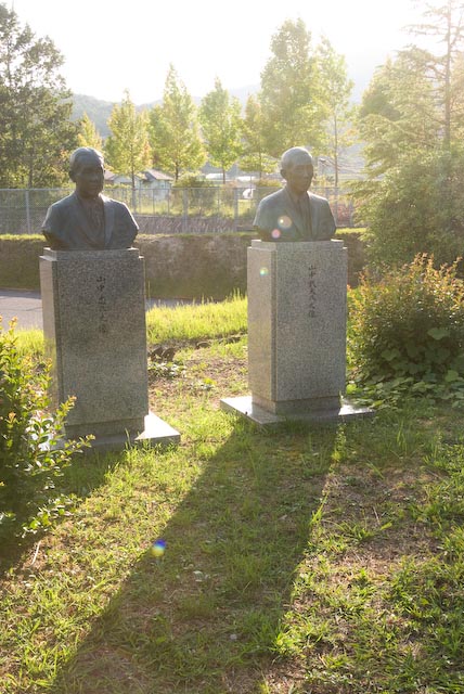 Two bronze statues of Takeo and Tadashi Yamanaka on stone pedestals in a garden setting with trees and hills in the background.