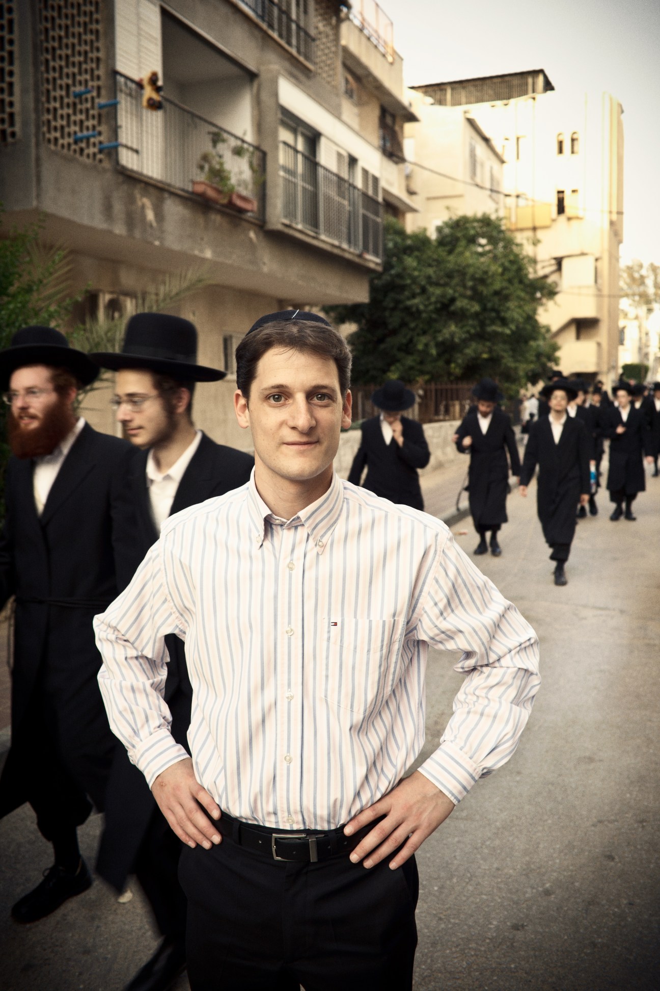 Eitan Dobkin stands in a striped shirt on a street in Bnei Brak, with Orthodox Jewish men in traditional black attire in the background.