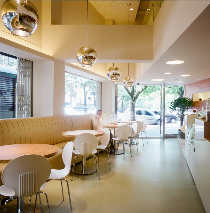 Modern café interior with yellow curved booth seating, white chairs, brass pendant lights, and street views.