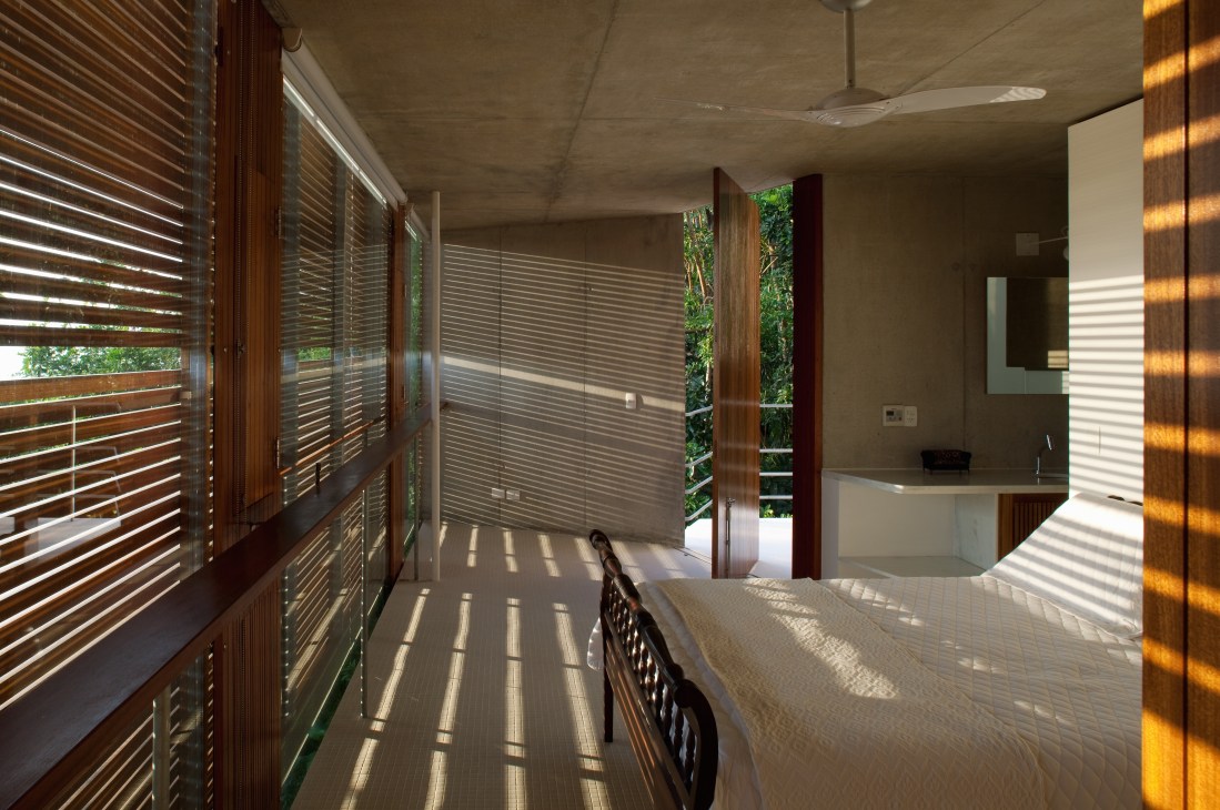 Main bedroom with wooden blinds filtering sunlight across concrete floor and bed.