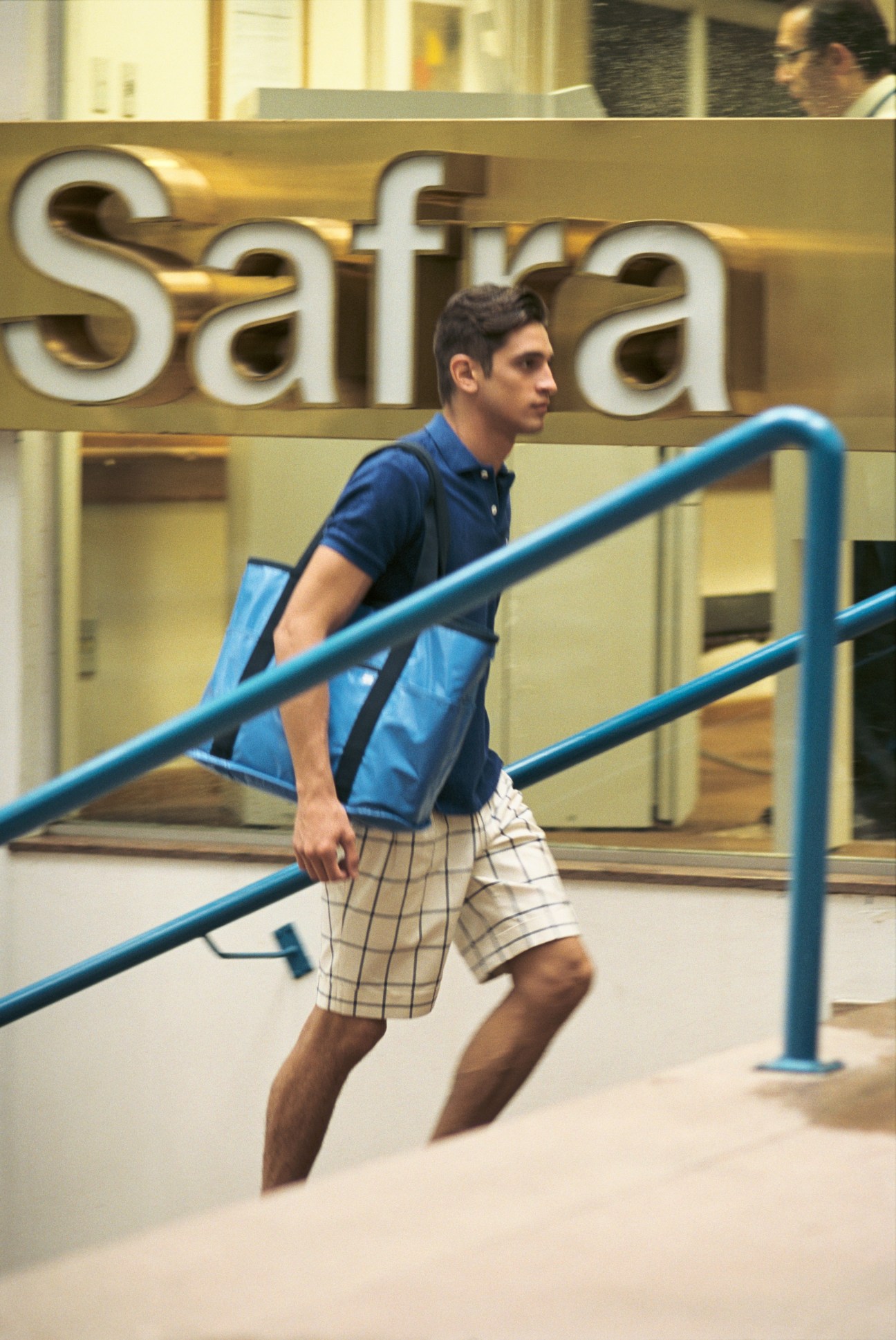 Model in blue polo shirt and plaid shorts stands on stairs at Hebraica sports club.