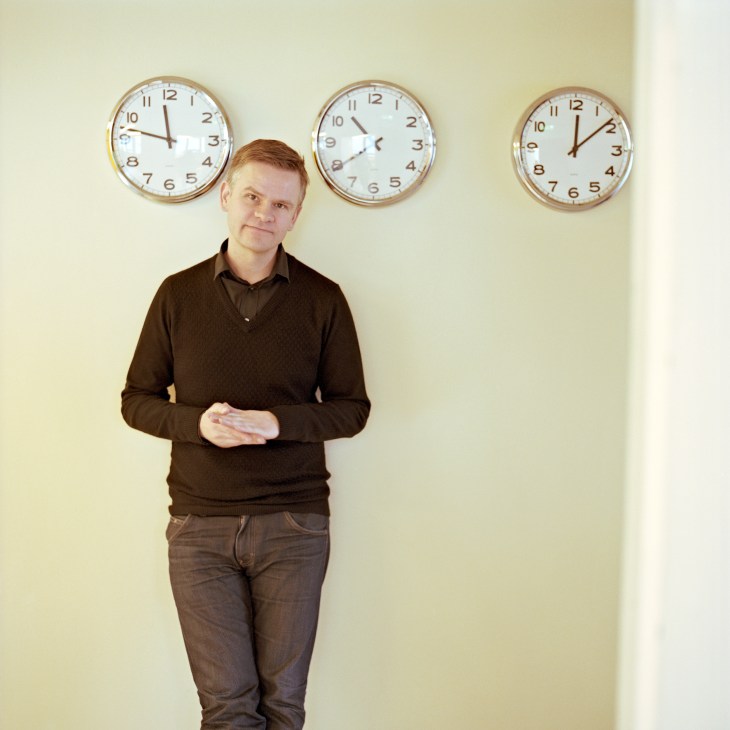 Man in brown shirt standing against pale wall with three clocks showing different times behind him.
