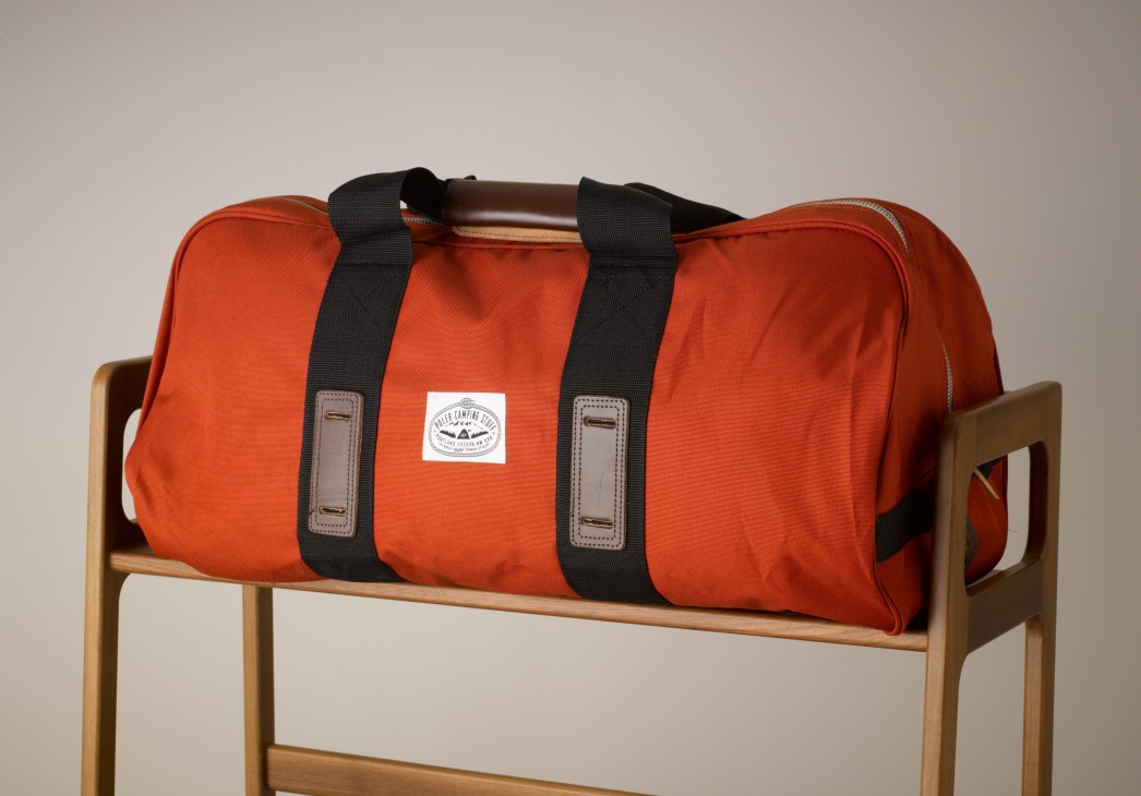 Poler Duffaluffagus duffle bag in rust-orange with black straps on wooden stand.