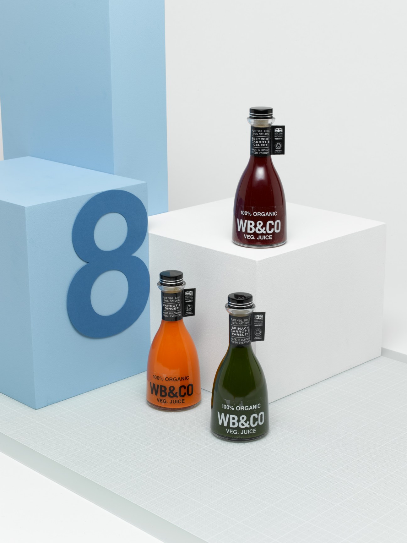 Three bottles of WB&Co organic vegetable juice in red, orange, and green displayed on white platforms next to a large blue number 8.