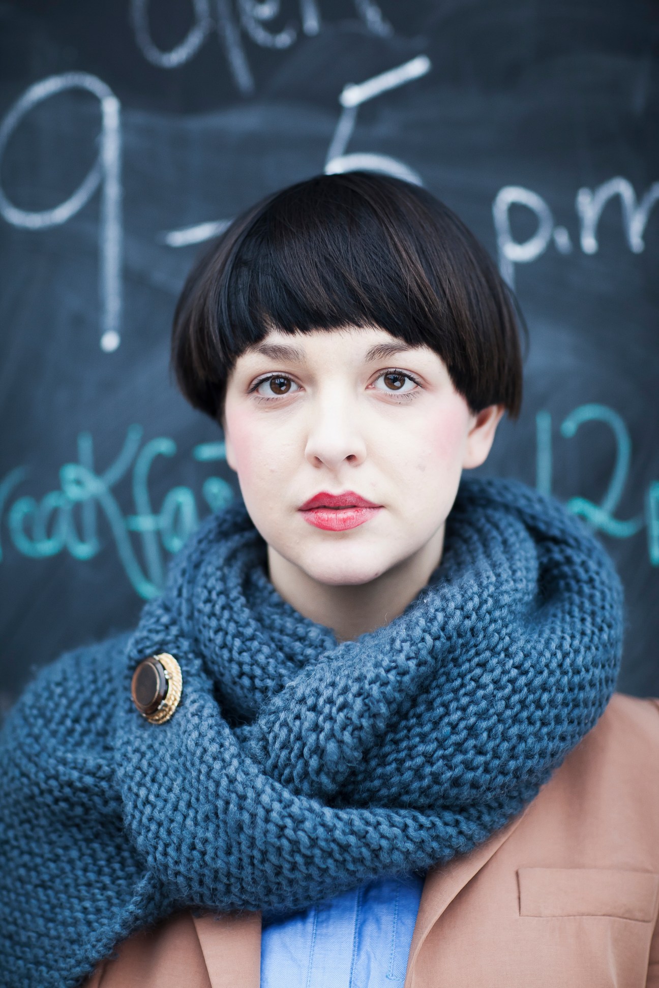 Woman with black bob haircut wearing blue knit scarf and gold brooch against chalkboard background.