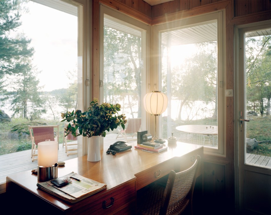 Wooden desk with lamp and flowers overlooking a terrace and water through large windows.