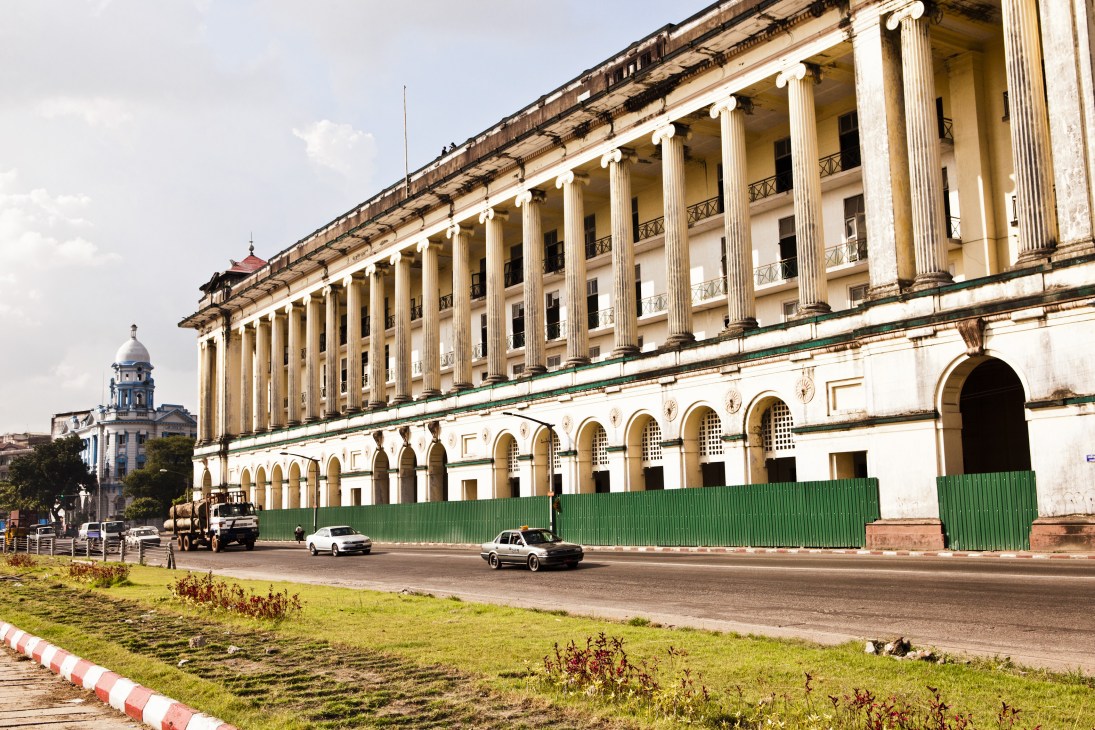 Colonial-era building with white neoclassical columns and arched windows, partially enclosed by green corrugated metal fencing.