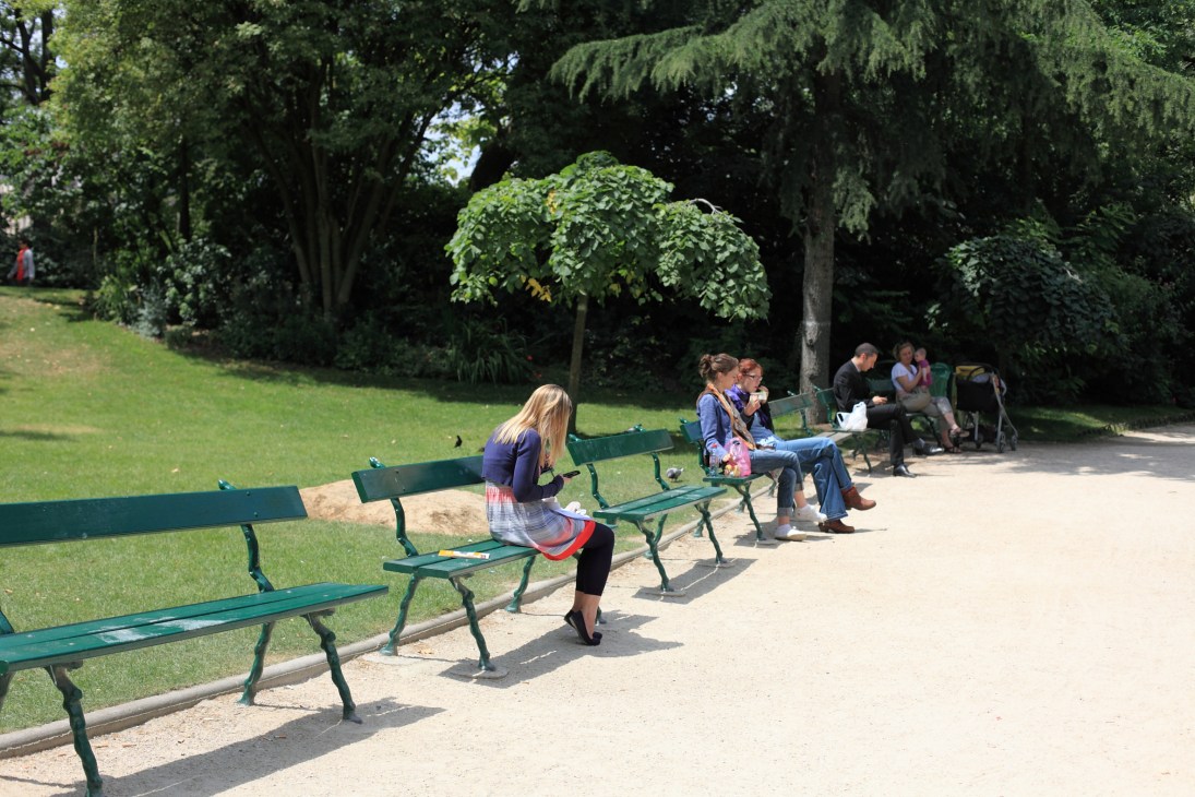 People sitting on green benches in a tree-lined park with manicured lawn.