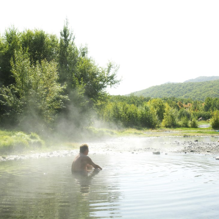 A person bathing in a steaming thermal pool surrounded by green forest and mountains in Kamchatka.