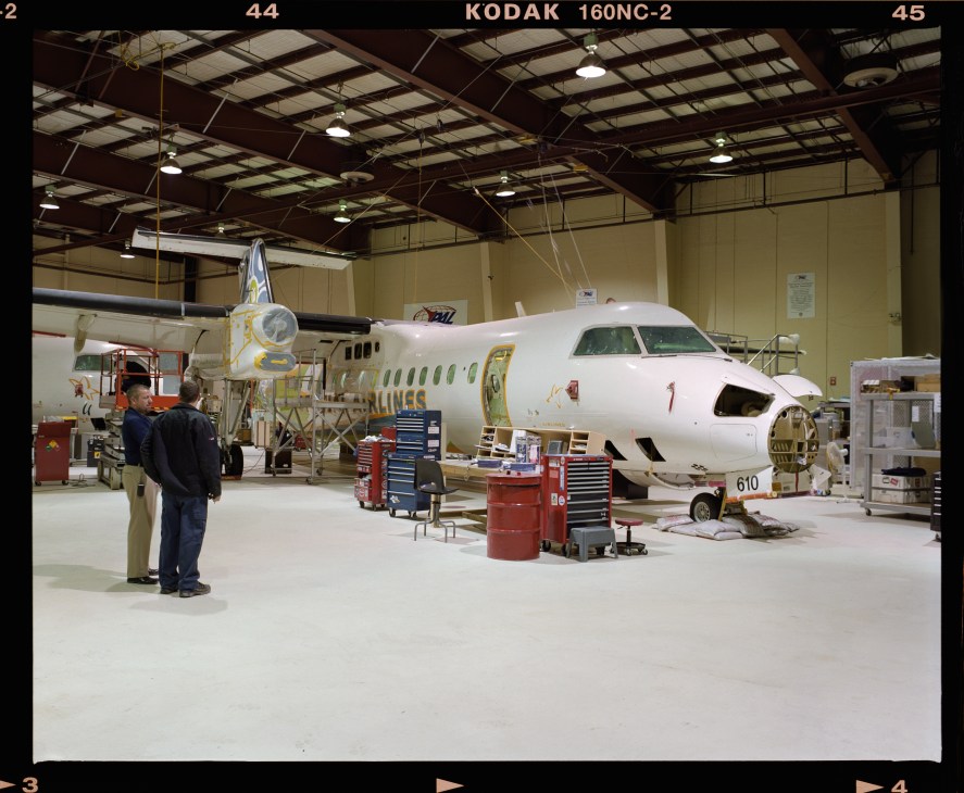 A white aircraft under maintenance in a large industrial hangar with workers and equipment visible.