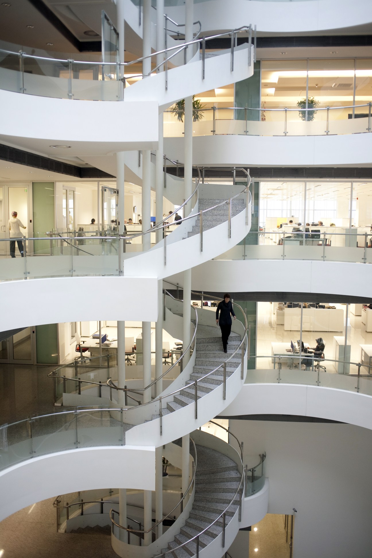 Interior of a modern multi-story office building with spiral staircase, glass railings, and workers on various floors.