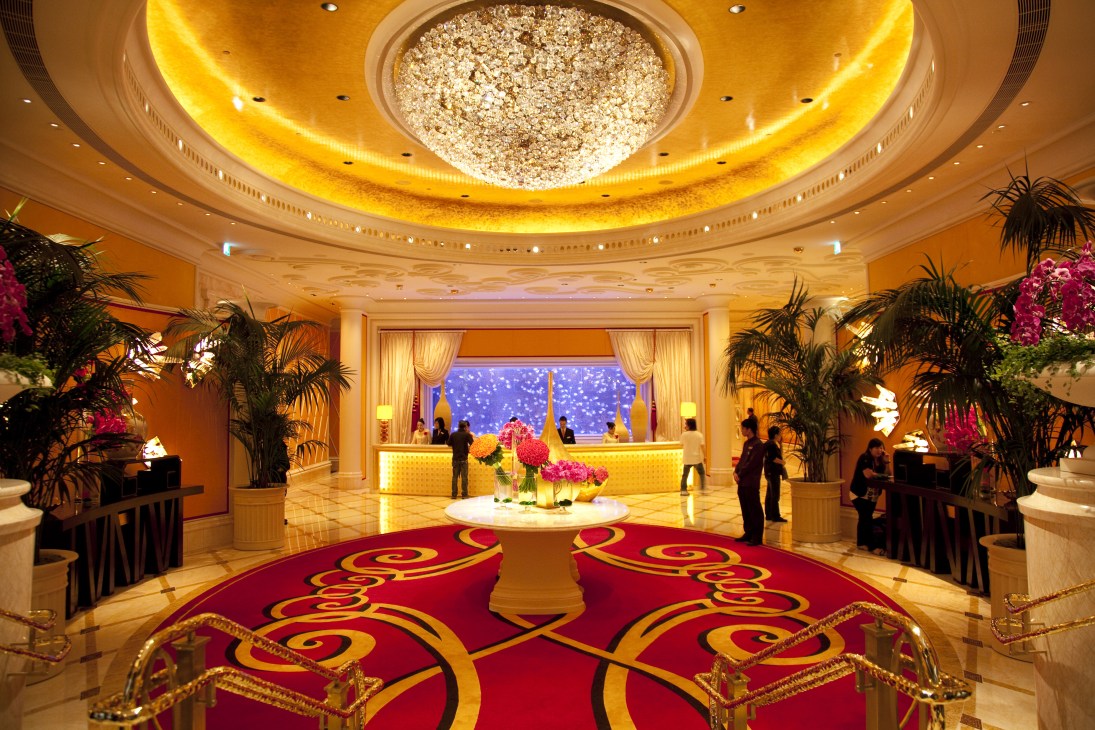 Luxurious hotel lobby with ornate chandelier, golden ceiling, red patterned carpet, and tropical plants at Wynn Resort Macau.