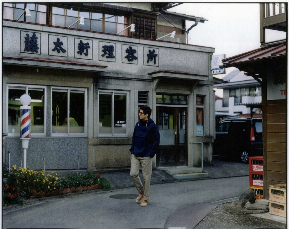 Man in glasses and Mackintosh coat stands outside Totaken barber shop in Tokyo suburb.