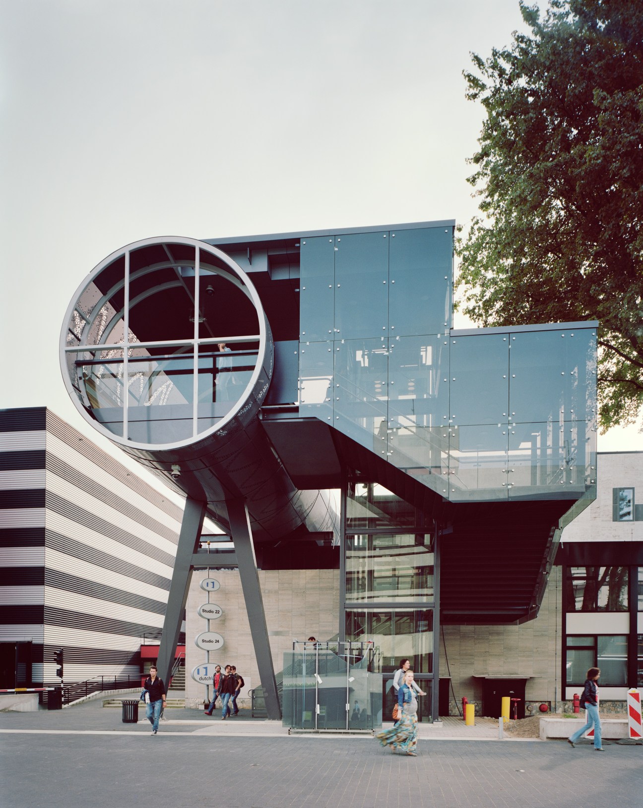 Modern architectural structure with large circular window and blue glass panels at Hilversum Noord station, with pedestrians walking nearby.