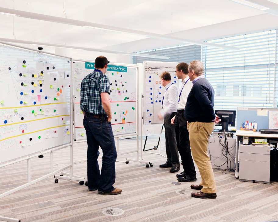 Four men discuss a project around whiteboards covered with notes and colored dots in a bright office.