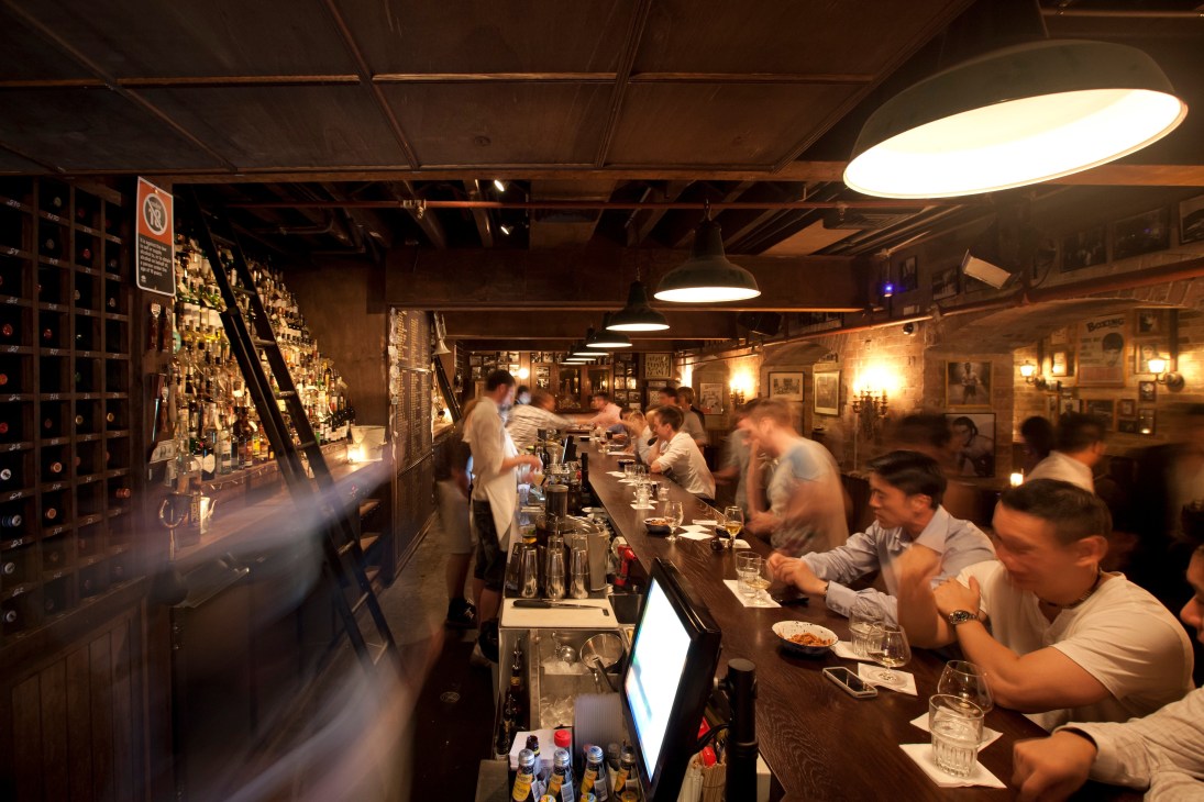 Interior of The Baxter Inn bar with patrons seated at counter and extensive whiskey selection on walls.