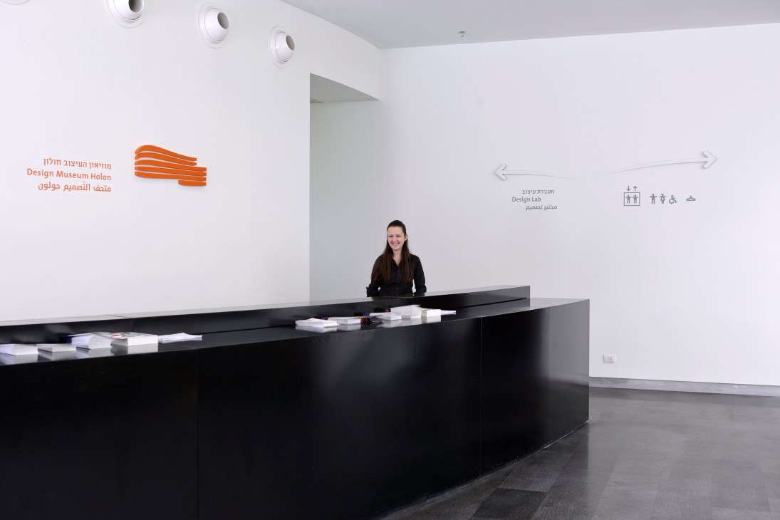 Design Museum Holon lobby with female staff member at black reception desk and wayfinding signage on white walls.