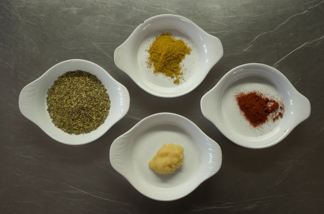 Four white bowls containing paprika, herbes de provence, garlic, and coriander on a dark surface.