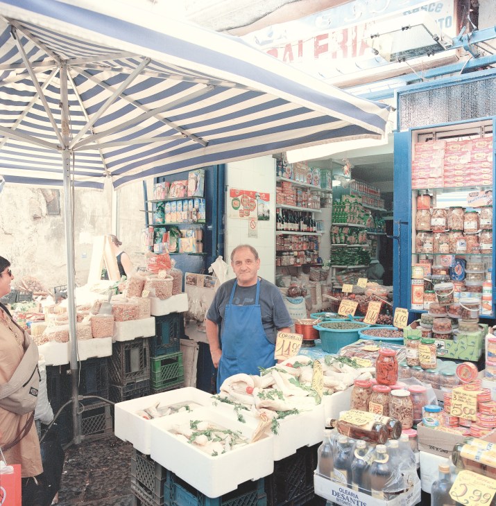 Stallholder in blue apron at Antignano Market in Naples, surrounded by packaged cured meats and food products.