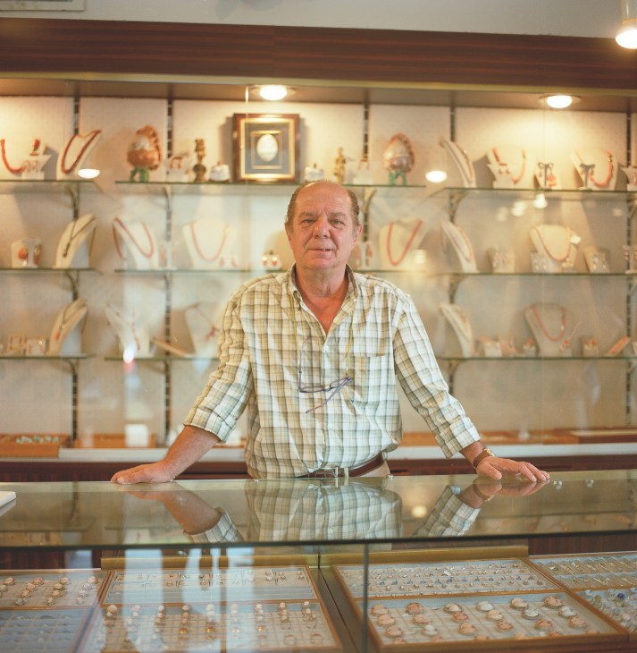 Antonio De Paola stands behind a display counter at his cameo factory, with shelves of jewelry and carved cameos visible behind him.