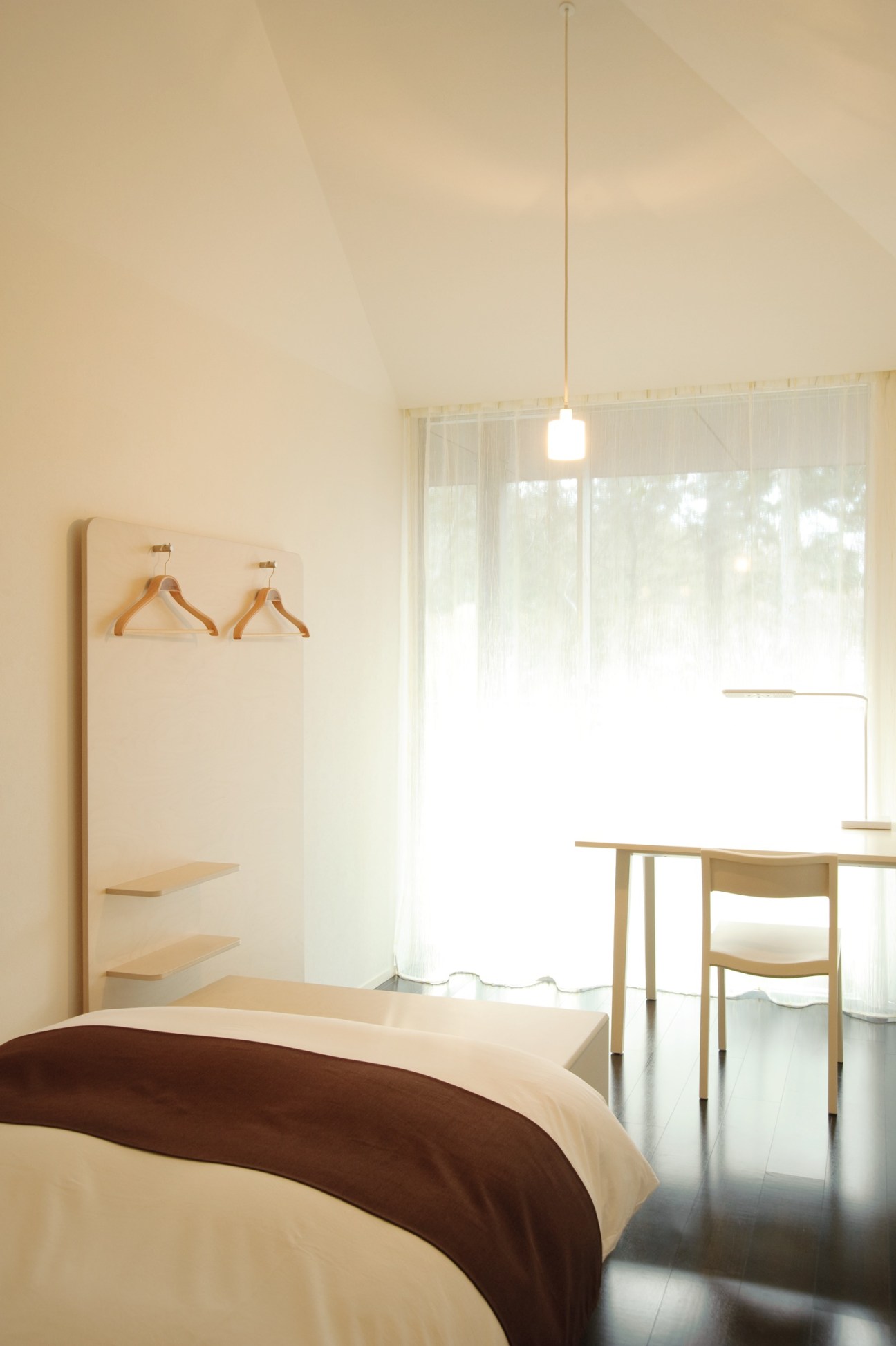 Minimalist bedroom with white walls, wooden desk, pendant light, and dark brown bedding with forest views through large windows.