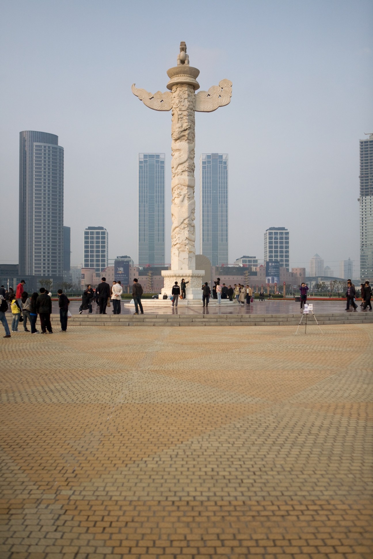 Ornate stone pillar with winged sculpture at top in large public square with visitors and modern buildings.