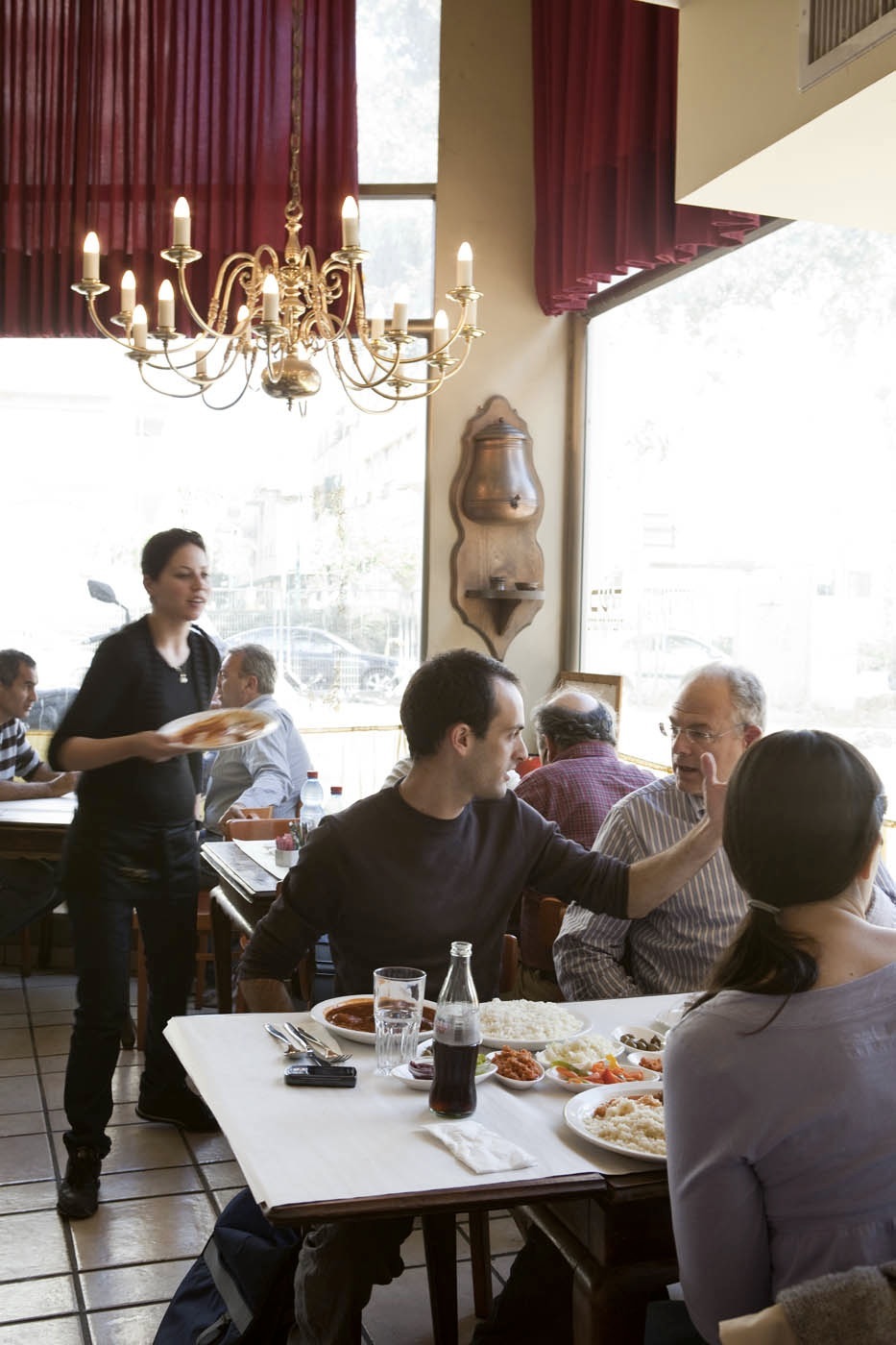 Diners enjoy lunch service at Gueta restaurant with a waitress serving plates beneath a brass chandelier.